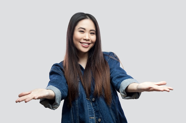 Portrait d'une belle jeune femme asiatique brune heureuse en veste bleue avec maquillage debout regardant la caméra avec les bras levés pour partager et sourire à pleines dents. tourné en studio, isolé sur fond gris clair.