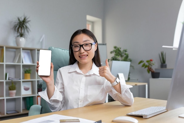 Portrait d'une belle jeune femme asiatique dans des verres tenant un téléphone portable dans sa main montrant le