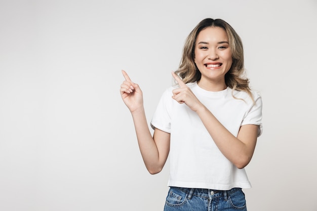 Portrait d'une belle jeune femme asiatique excitée debout isolée sur un mur blanc, pointant vers l'espace de copie