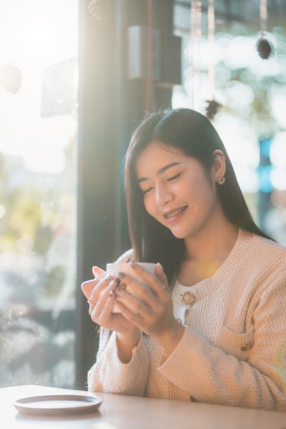 Portrait belle jeune femme asiatique fille portant des vêtements chauds aime boire en tenant positif la tasse de café, de lait ou de chocolat à la maison dans le salon à l'intérieur ou au café