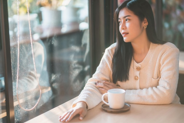 Portrait belle jeune femme asiatique fille portant des vêtements chauds aime boire en tenant positif la tasse de café, de lait ou de chocolat à la maison dans le salon à l'intérieur ou au café