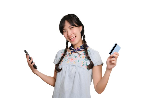 Portrait d'une belle jeune femme asiatique heureuse en robe denim tenant une carte de crédit et un smartphone sur fond blanc tourné en studio