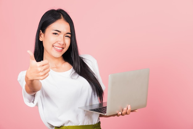 Portrait d'une belle jeune femme asiatique heureuse visage souriant confiant tenant à l'aide d'un ordinateur portable et montrant le pouce vers le haut pour un geste similaire, prise de vue en studio avec espace de copie isolé sur fond rose