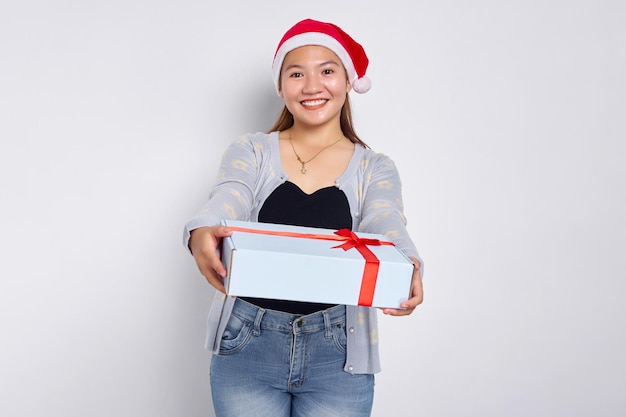 Portrait d'une belle jeune femme asiatique souriante portant un chandail bleu avec un chapeau de père Noël donnant une boîte-cadeau isolée sur fond de studio blanc Joyeux Noël Concept