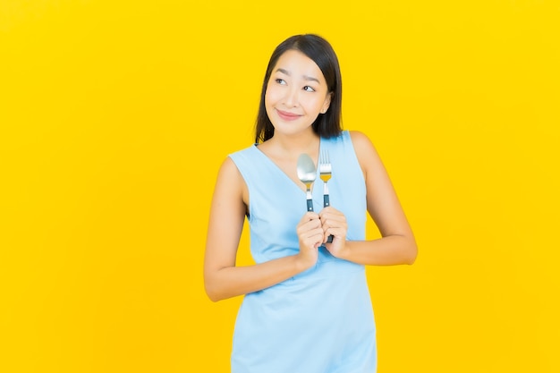 Portrait belle jeune femme asiatique sourire avec cuillère et fourchette sur le mur de couleur jaune