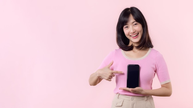 Photo portrait belle jeune femme asiatique sourire heureux montrant l'écran du smartphone avec le geste de la main du doigt pointé vers le téléphone mobile isolé sur fond de studio pastel rose concept de smartphone à écran blanc