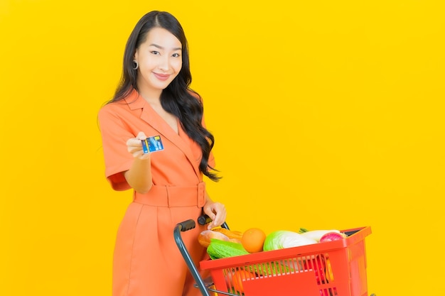 Portrait belle jeune femme asiatique sourire avec panier d'épicerie de supermarché sur jaune