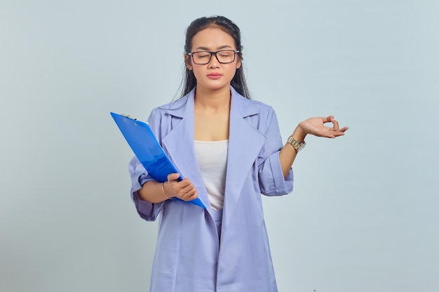 Portrait d'une belle jeune femme asiatique tenant un dossier de documents et debout dans la pose de lotus, essayant de méditer le yoga, fermer les yeux isolés sur fond violet