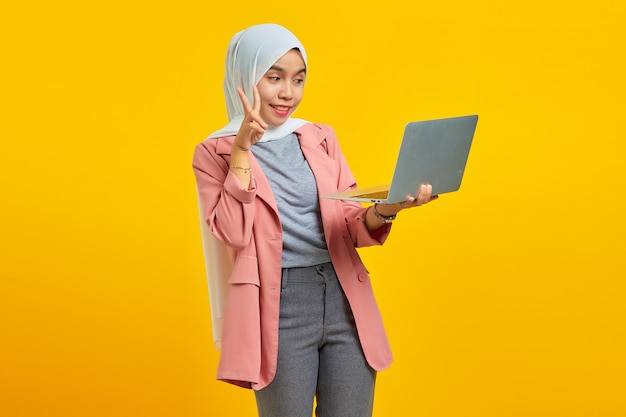 Portrait d'une belle jeune femme asiatique tenant un ordinateur portable et faisant un signe de paix avec les doigts isolés sur fond jaune