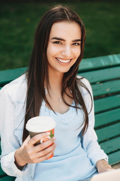 Portrait d'une belle jeune femme assise sur un banc avec une tasse de café dans une main regardant la caméra en riant à l'extérieur dans la ville.