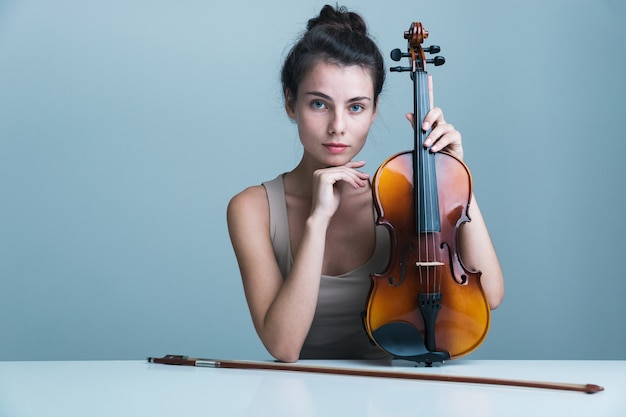 Portrait d'une belle jeune femme assise à la table avec un violon isolé sur fond bleu