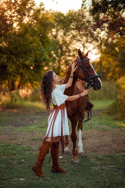 Portrait d'une belle jeune femme aux cheveux bouclés qui tient un cheval brun Marcher avec les animaux de la ferme Équitation cavalier de style boho