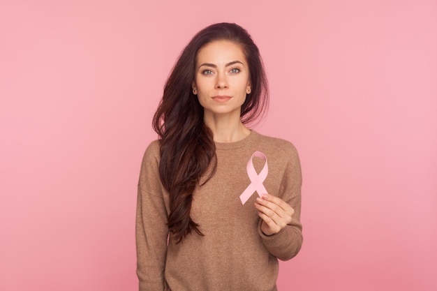 Portrait de la belle jeune femme aux cheveux bruns tenant un ruban rose symbole du soutien du cancer du sein des patients en oncologie tourné en studio isolé sur fond rose