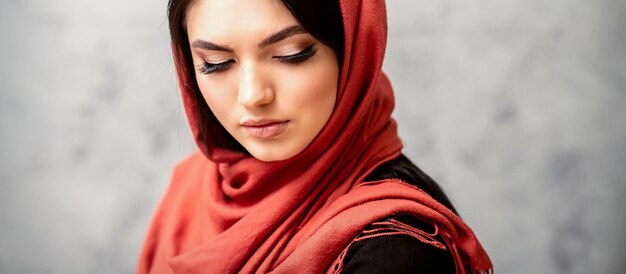 Portrait de la belle jeune femme aux cheveux longs et maquillage avec des extensions de cils dans une écharpe rouge Beauté jeune femme caucasienne sur le fond d'un mur gris