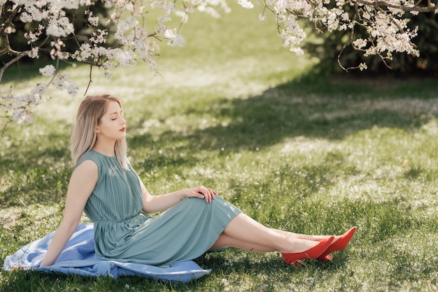 Portrait d'une belle jeune femme blonde assise sous un arbre en fleurs dans le parc et appréciant la chaleur