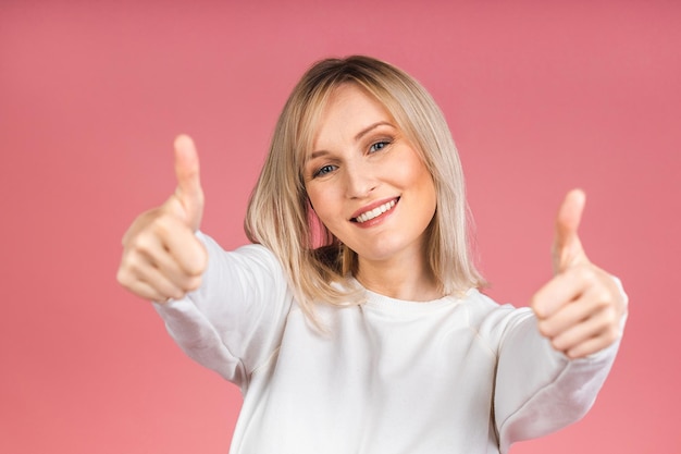 Portrait d'une belle jeune femme blonde joyeuse mignonne souriante regardant la caméra isolée sur fond rose. Signe pouce vers le haut.