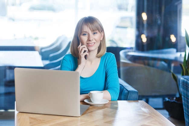 Portrait de la belle jeune femme blonde en t-shirt bleu assis et travaillant au café avec ordinateur portable. tenant un téléphone portable, parler et regarder la caméra avec un sourire à pleines dents. intérieur, prise de vue en studio.