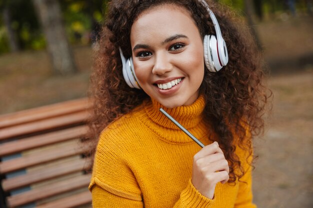 Portrait d'une belle jeune femme bouclée souriante assise sur un banc dans un parc à l'extérieur, écoutant de la musique avec un casque tenant un crayon.