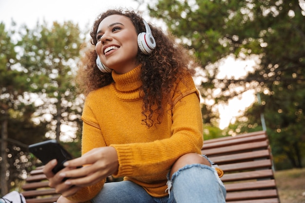 Portrait d'une belle jeune femme bouclée souriante heureuse s'asseoir sur un banc dans un parc à l'extérieur, écouter de la musique avec des écouteurs à l'aide d'un téléphone portable.