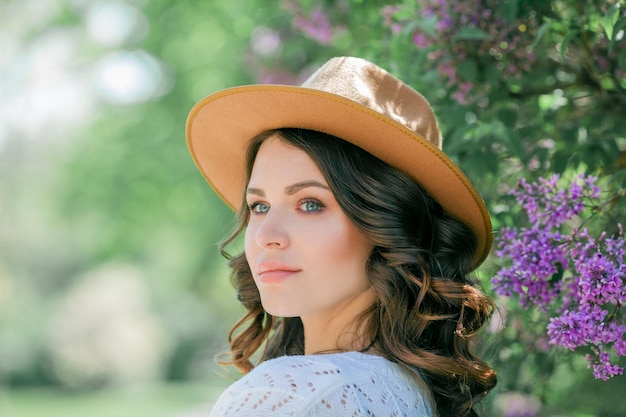 Portrait de la belle jeune femme brune aux yeux bleus en chapeau près du modèle heureux en fleurs avec des cheveux bouclés printemps