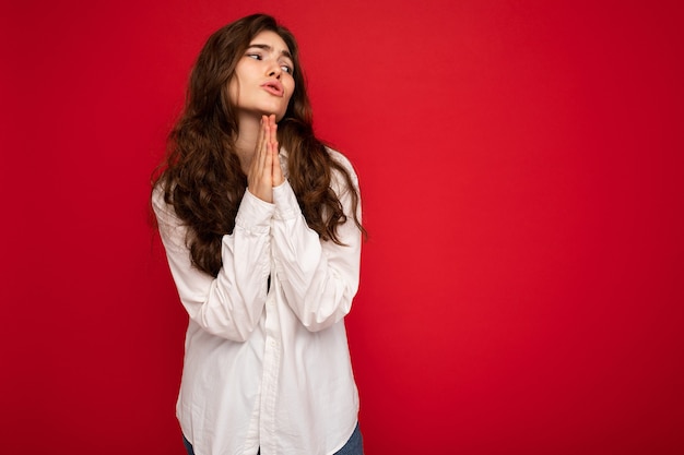 Portrait de belle jeune femme brune bouclée vêtue d'une chemise blanche isolée sur fond rouge avec