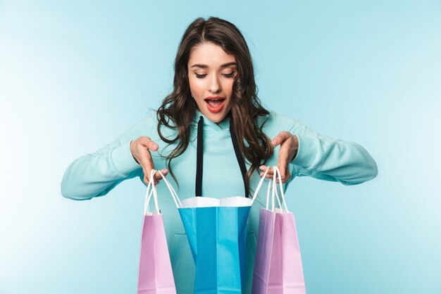 Portrait d'une belle jeune femme brune excitée debout sur bleu, portant des sacs à provisions