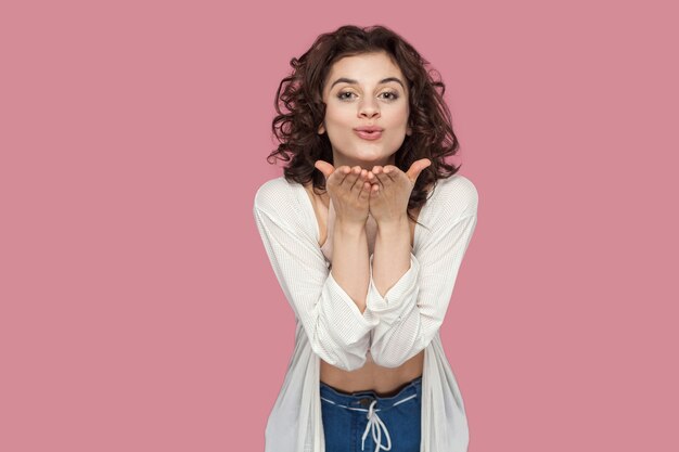 Portrait D'une Belle Jeune Femme Brune Heureuse En Amour Avec Une Coiffure Frisée Dans Un Style Décontracté, Debout Et Envoyant Un Baiser Aérien Et Regardant La Caméra. Tourné En Studio Intérieur Isolé Sur Fond Rose.