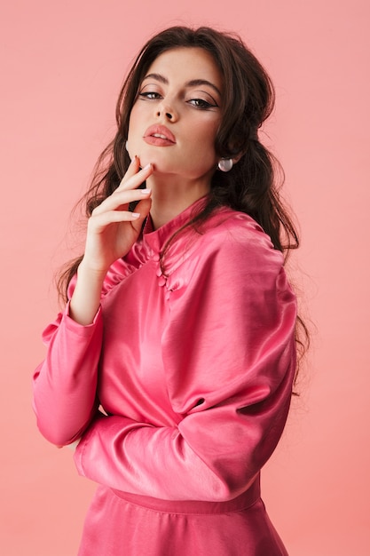 Portrait d'une belle jeune femme brune avec un maquillage lumineux portant des vêtements à la mode debout isolé sur un mur rose, posant