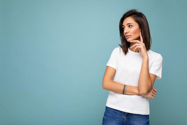 Portrait d'une belle jeune femme brune réfléchie avec des émotions sincères portant un t-shirt blanc décontracté pour une maquette isolée sur fond bleu avec espace de copie et réflexion.