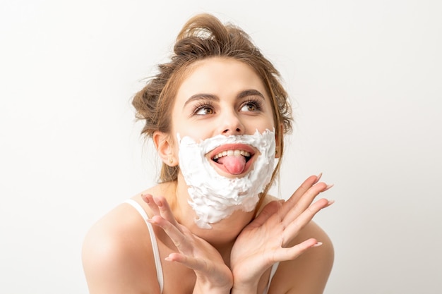 Portrait d'une belle jeune femme caucasienne souriante posant avec de la mousse à raser sur le visage sorti sa langue sur fond blanc.