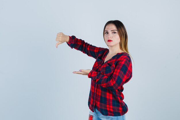 Portrait d'une belle jeune femme écartant la paume de la main, montrant le pouce vers le bas en chemise décontractée et regardant la vue de face sans gaieté