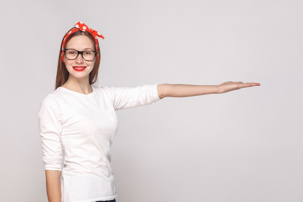 Portrait d'une belle jeune femme émotive en t-shirt blanc avec des lunettes, des lèvres rouges et un bandeau regardant la caméra avec un sourire à pleines dents et montrant un espace de copie. tir à l'intérieur, isolé sur fond gris.