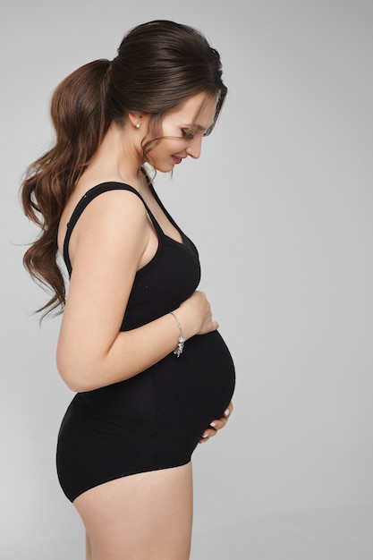 Portrait d'une belle jeune femme enceinte en body noir posant sur fond gris isolé ...