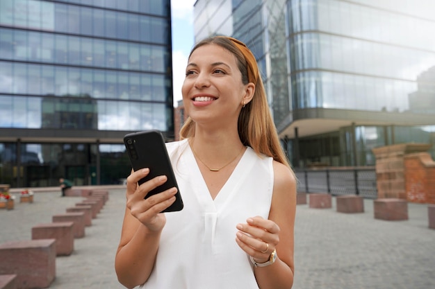 Portrait d'une belle jeune femme entrepreneur regardant loin tenant un smartphone contre des bâtiments commerciaux Charmante jeune femme allant travailler