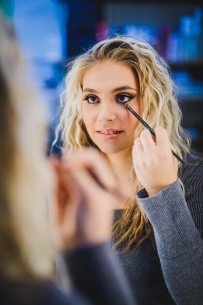 Portrait d'une belle jeune femme faisant du maquillage en regardant dans le miroir et en appliquant un eye-liner avec un pinceau.