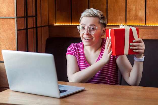 Portrait d'une belle jeune femme heureuse aux cheveux courts en t-shirt rose et lunettes est assise dans un café, tenant une boîte cadeau rouge, facile à commander en ligne, regardant la caméra et souriant à pleines dents. Intérieur