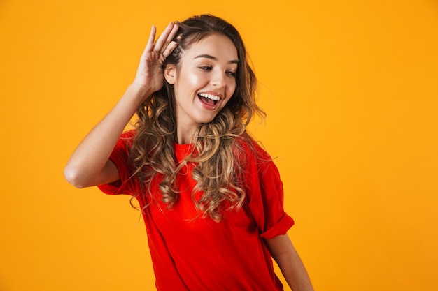 Portrait D'une Belle Jeune Femme Joyeuse Debout Isolée Sur Un Mur Jaune, Essayant D'entendre Des Rumeurs