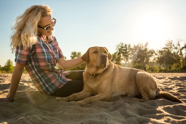 Portrait d'une belle jeune femme à lunettes de soleil assise sur une plage de sable avec une fille de chien golden retriever...