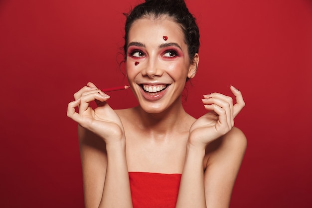 Portrait d'une belle jeune femme avec un maquillage rouge vif isolé sur un mur rouge posant avec des coeurs sur le visage tenant un crayon à lèvres.