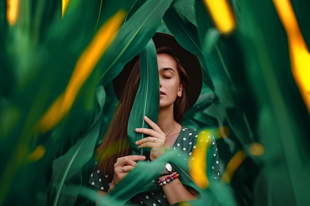 Portrait d'une belle jeune femme à la mode sensuelle et élégante avec une peau de visage lisse, des yeux fermés et un maquillage naturel portant un chapeau et une robe à pois tenant une feuille verte dans ses mains