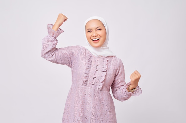 Portrait d'une belle jeune femme musulmane asiatique excitée portant le hijab et une robe violette levant les poings et célébrant la victoire du Ramadan isolée sur fond de studio blanc