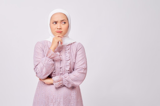 Portrait d'une belle jeune femme musulmane asiatique pensive portant le hijab et une robe violette regardant de côté tout en touchant son menton en pensant à l'offre isolée sur fond de studio blanc