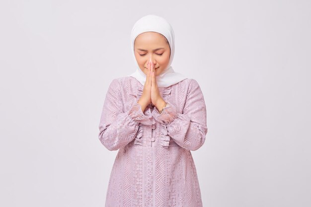 Portrait de la belle jeune femme musulmane asiatique portant le hijab et la robe violette debout avec la main priant Dieu et disant merci pour la faveur donnée isolé sur fond de studio blanc