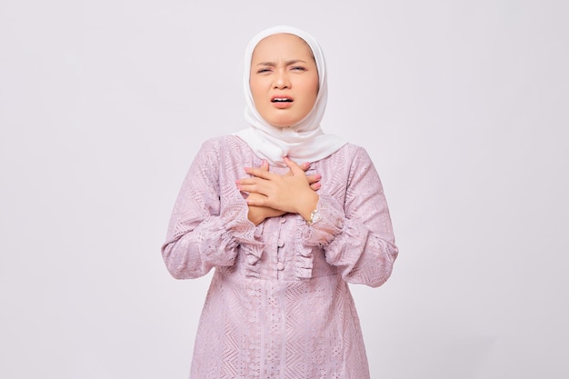 Portrait d'une belle jeune femme musulmane asiatique portant le hijab et une robe violette pressant la main sur la poitrine ayant une crise cardiaque isolée sur fond de studio blanc