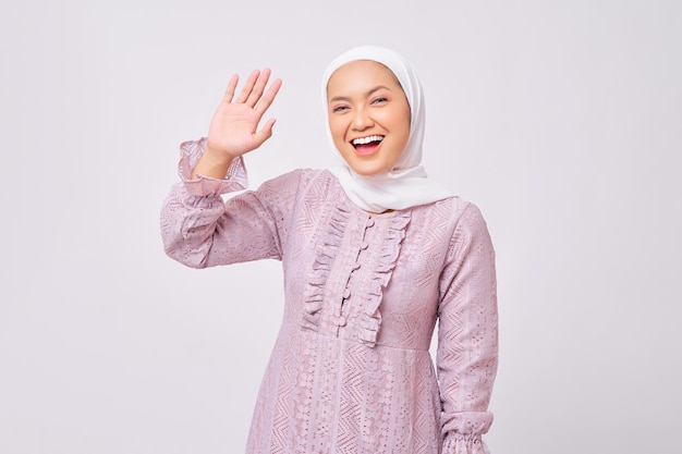 Portrait d'une belle jeune femme musulmane asiatique souriante portant le hijab et une robe violette dites bonjour à la caméra et agitant la main pour saluer quelqu'un isolé sur fond de studio blanc