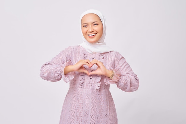 Portrait d'une belle jeune femme musulmane asiatique souriante portant le hijab et une robe violette montrant un geste de coeur faisant signe d'amour avec les doigts isolés sur fond de studio blanc