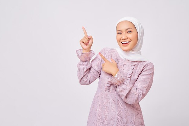 Portrait d'une belle jeune femme musulmane asiatique souriante portant le hijab et une robe violette pointant le doigt sur un espace libre pour une publicité isolée sur fond de studio blanc