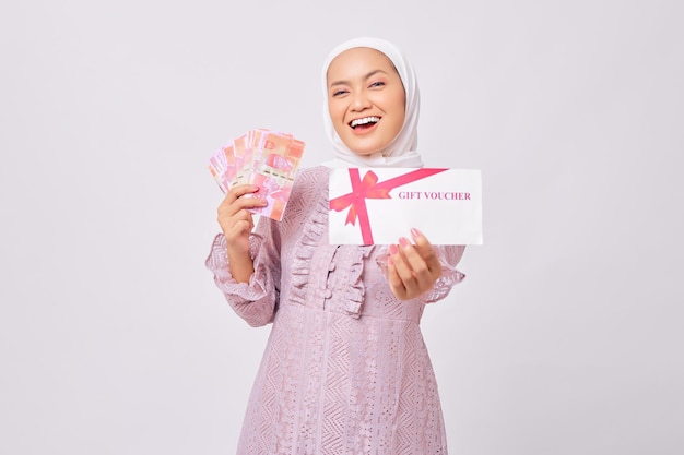 Portrait d'une belle jeune femme musulmane asiatique souriante portant le hijab et une robe violette tenant un certificat-cadeau et des billets en roupies en espèces isolés sur fond de studio blanc