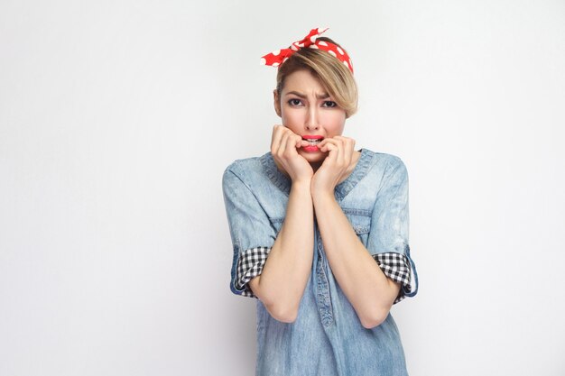 Portrait d'une belle jeune femme nerveuse en chemise en jean bleu décontractée avec maquillage et bandeau rouge debout regardant la caméra et se rongeant les ongles. tourné en studio intérieur, isolé sur fond blanc.