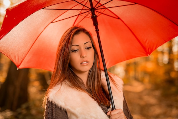 Portrait d'une belle jeune femme pensive avec un parapluie rouge dans une forêt ensoleillée aux couleurs de l'automne.